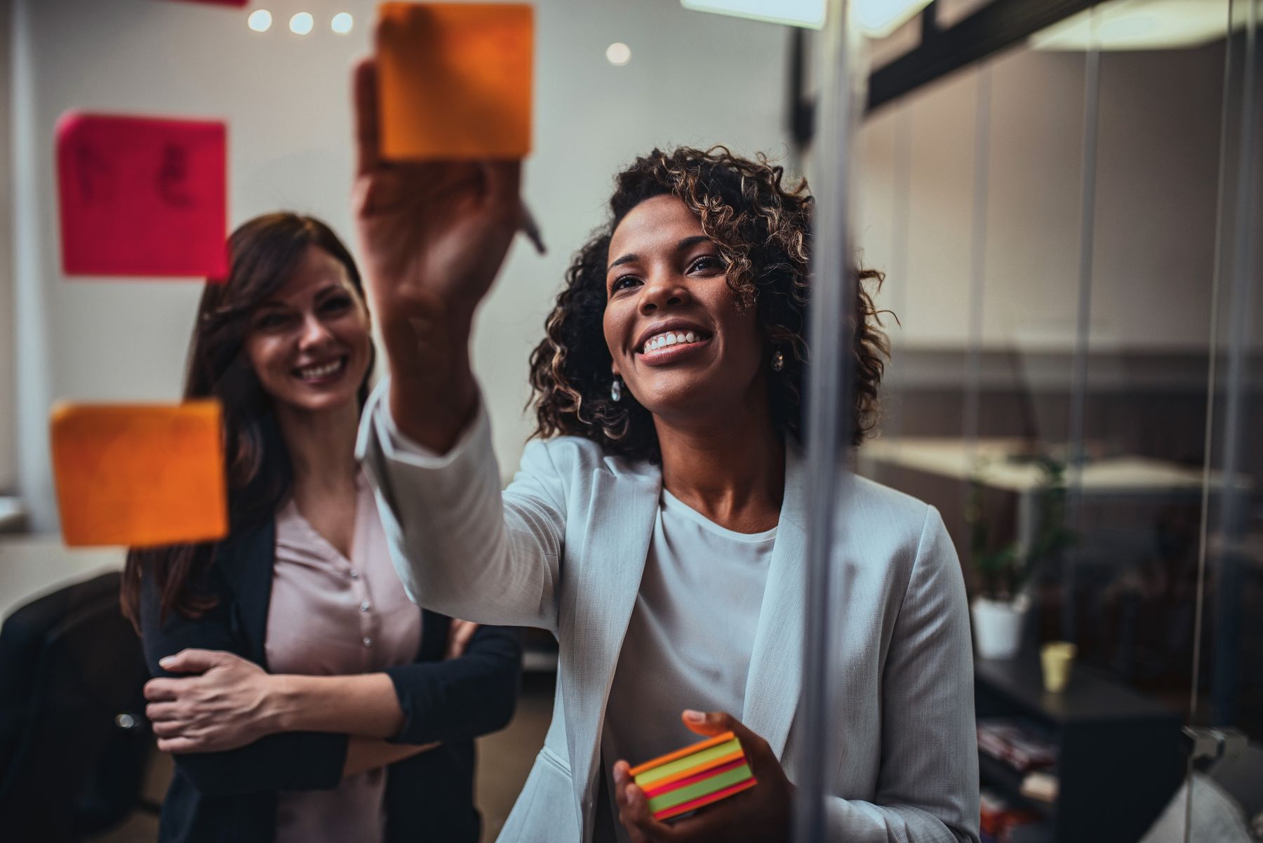 Duas colegas sorrindo diante de um quadro de vidro com notas adesivas